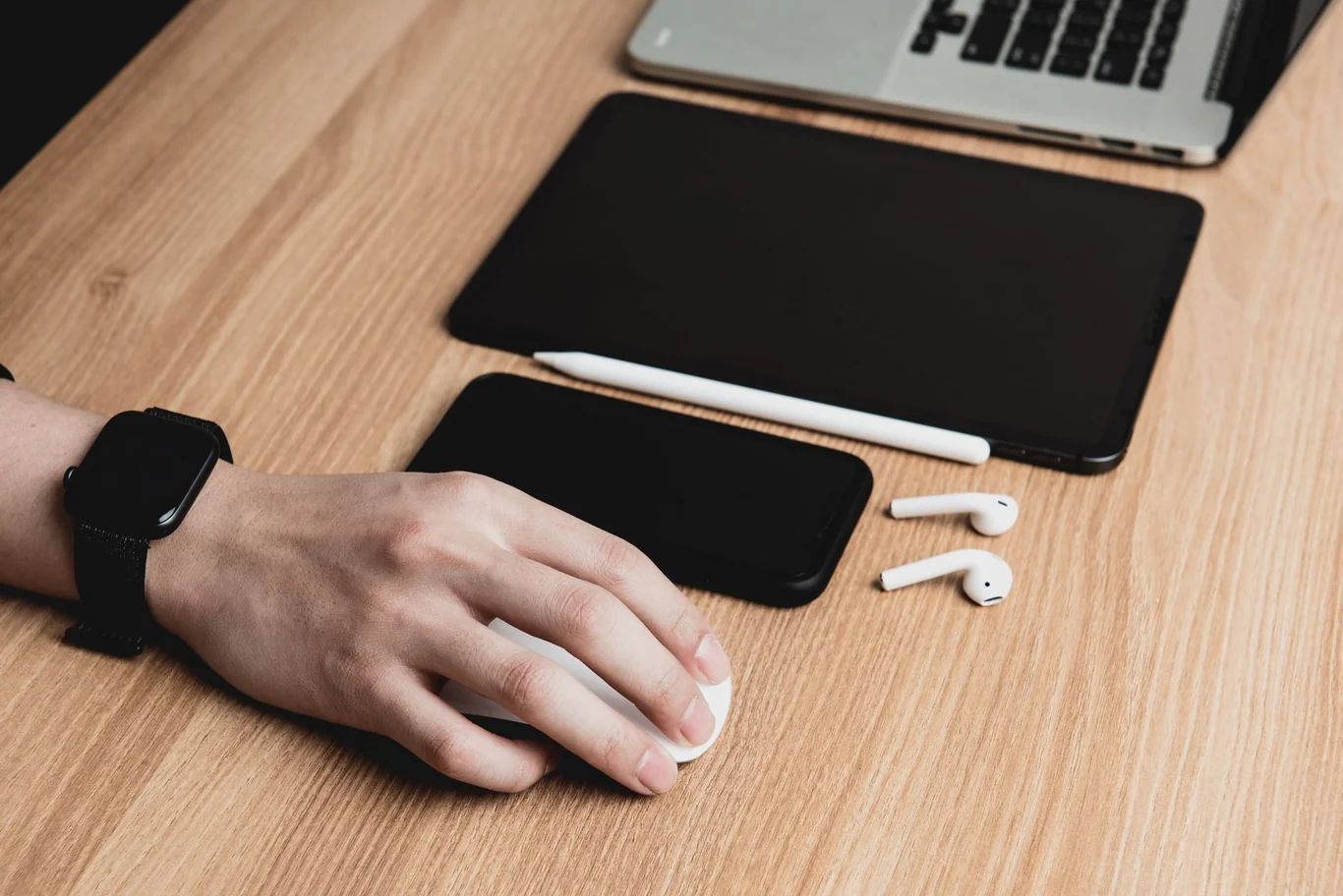 Black smartwatch on a wrist beside a laptop, tablet and wireless earbuds on a desk