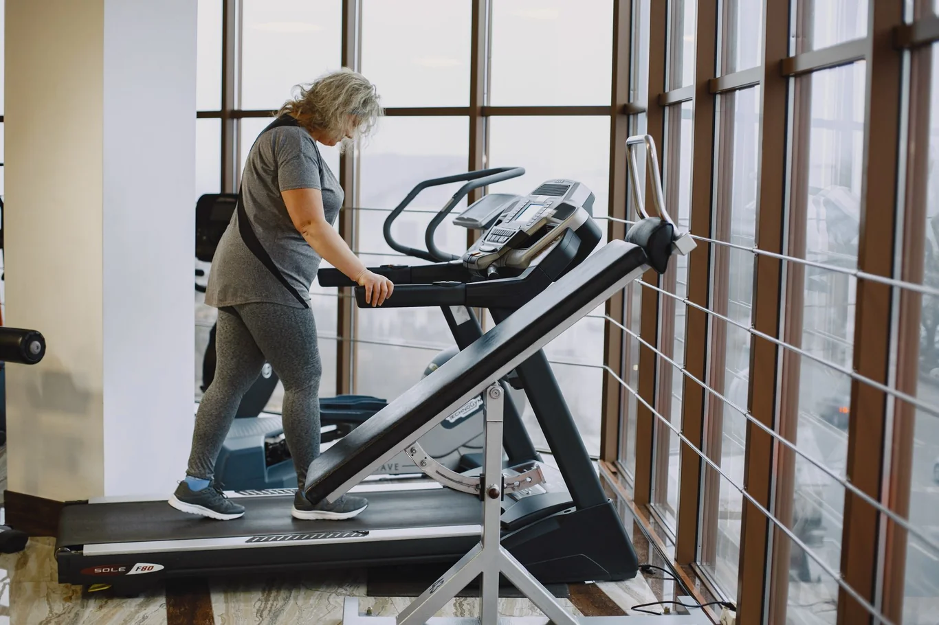 Older woman walking on an inclined treadmill in a gym near large windows