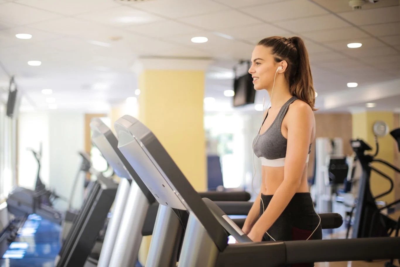 Woman in workout gear standing at a treadmill console adjusting settings in a gym