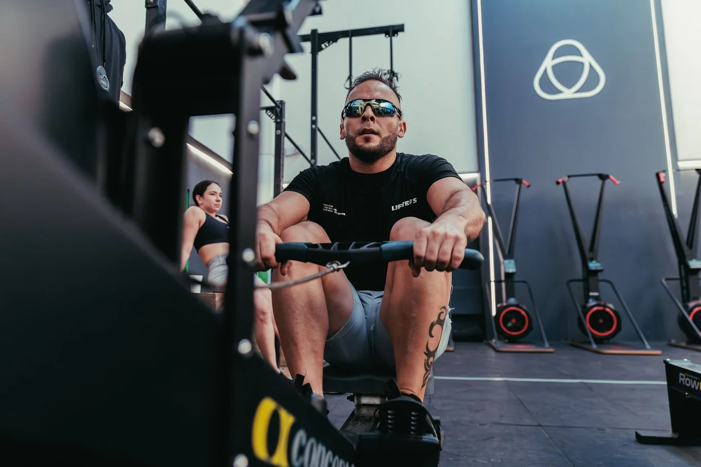 Man in sunglasses gripping the handle bar of a rowing machine mid-workout in a gym