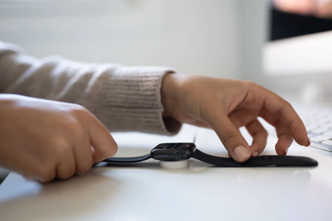 Hands attach a white charging cable to a black smartwatch on a desk