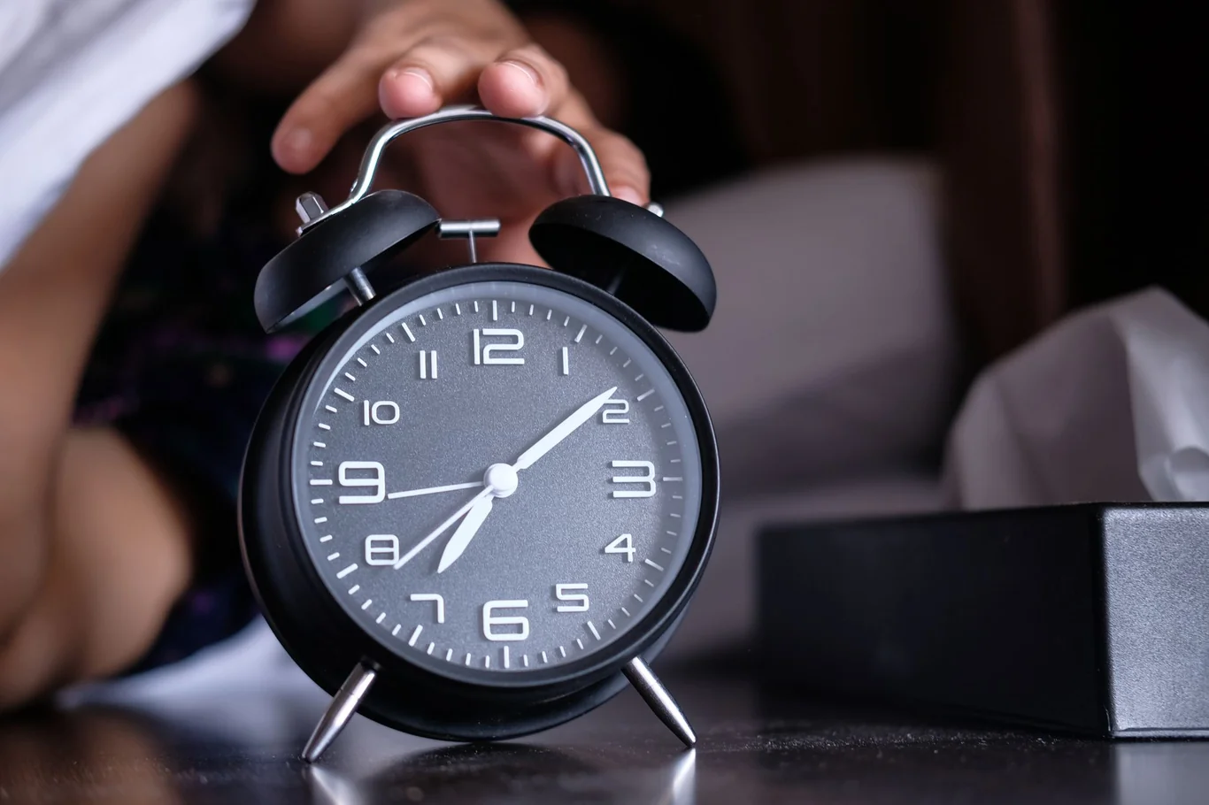 A hand reaching to turn off a black alarm clock beside a bed in the morning