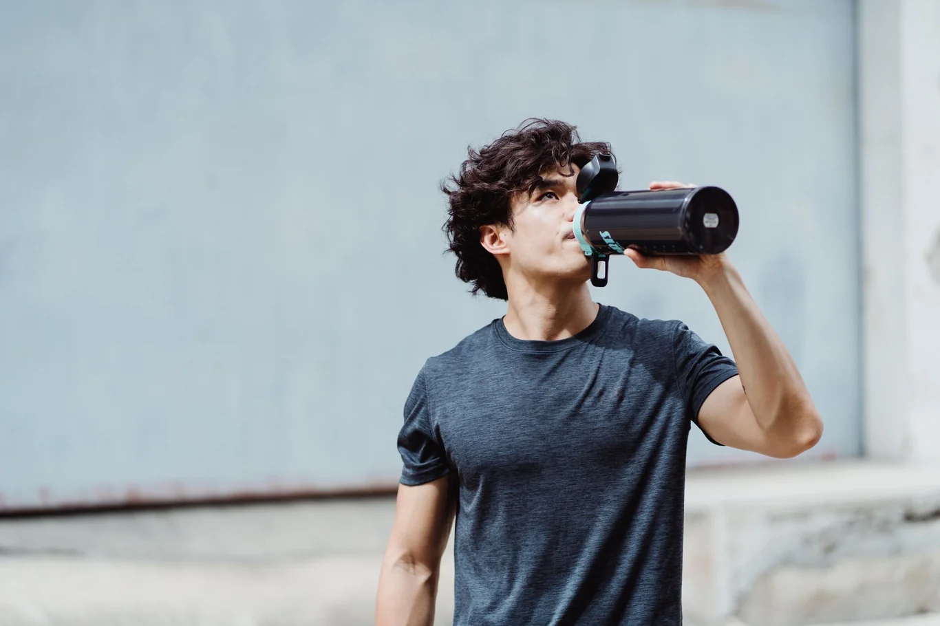 Young man drinking water from a shaker bottle outdoors against a plain wall