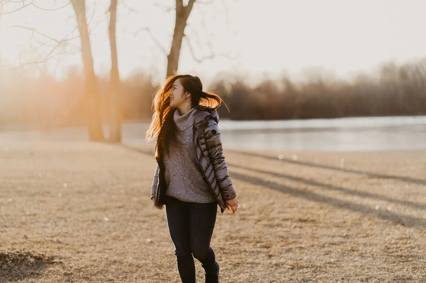 Woman walking outdoors near a lake at golden hour wearing a knit sweater and jacket