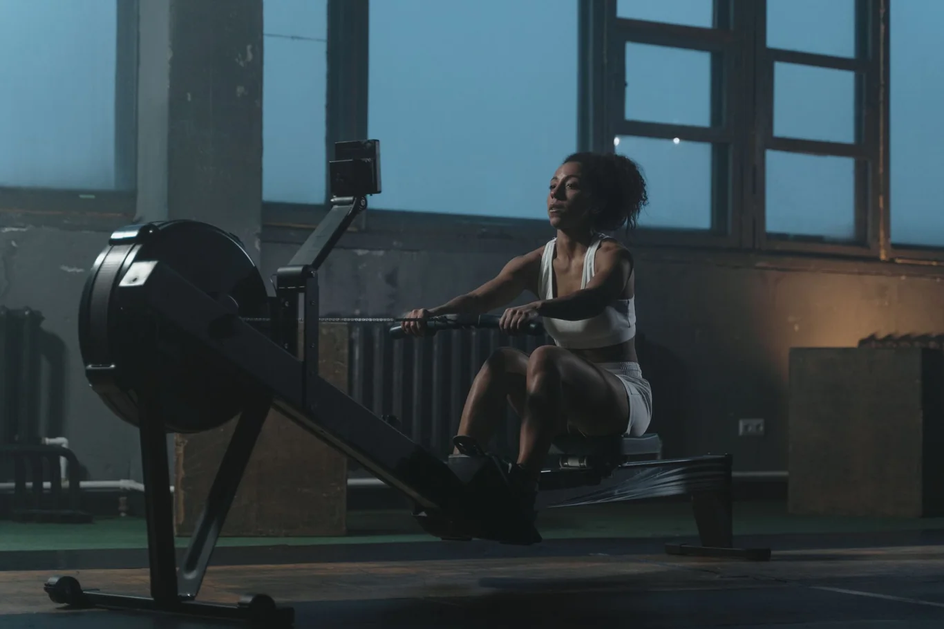 Woman rowing on an indoor rowing machine in a dark gym with window light
