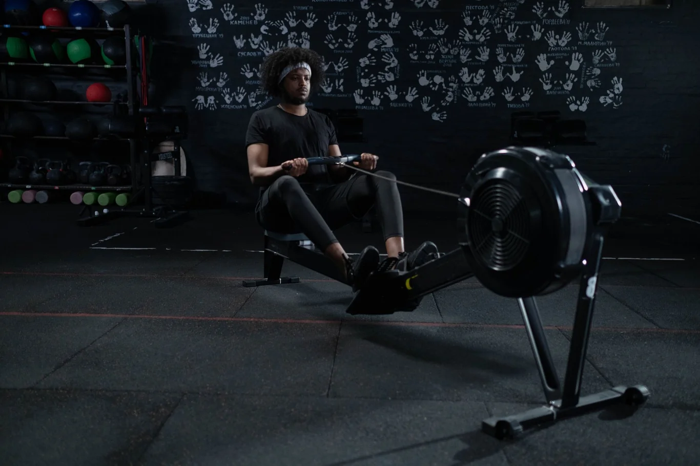 Man rowing on an indoor rowing machine with a flywheel fan cage in a dim gym