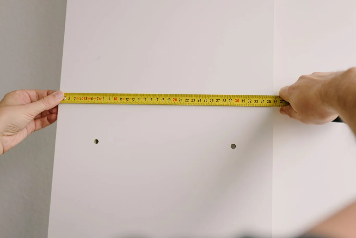 Two hands holding a yellow tape measure across a white wall with two drilled holes