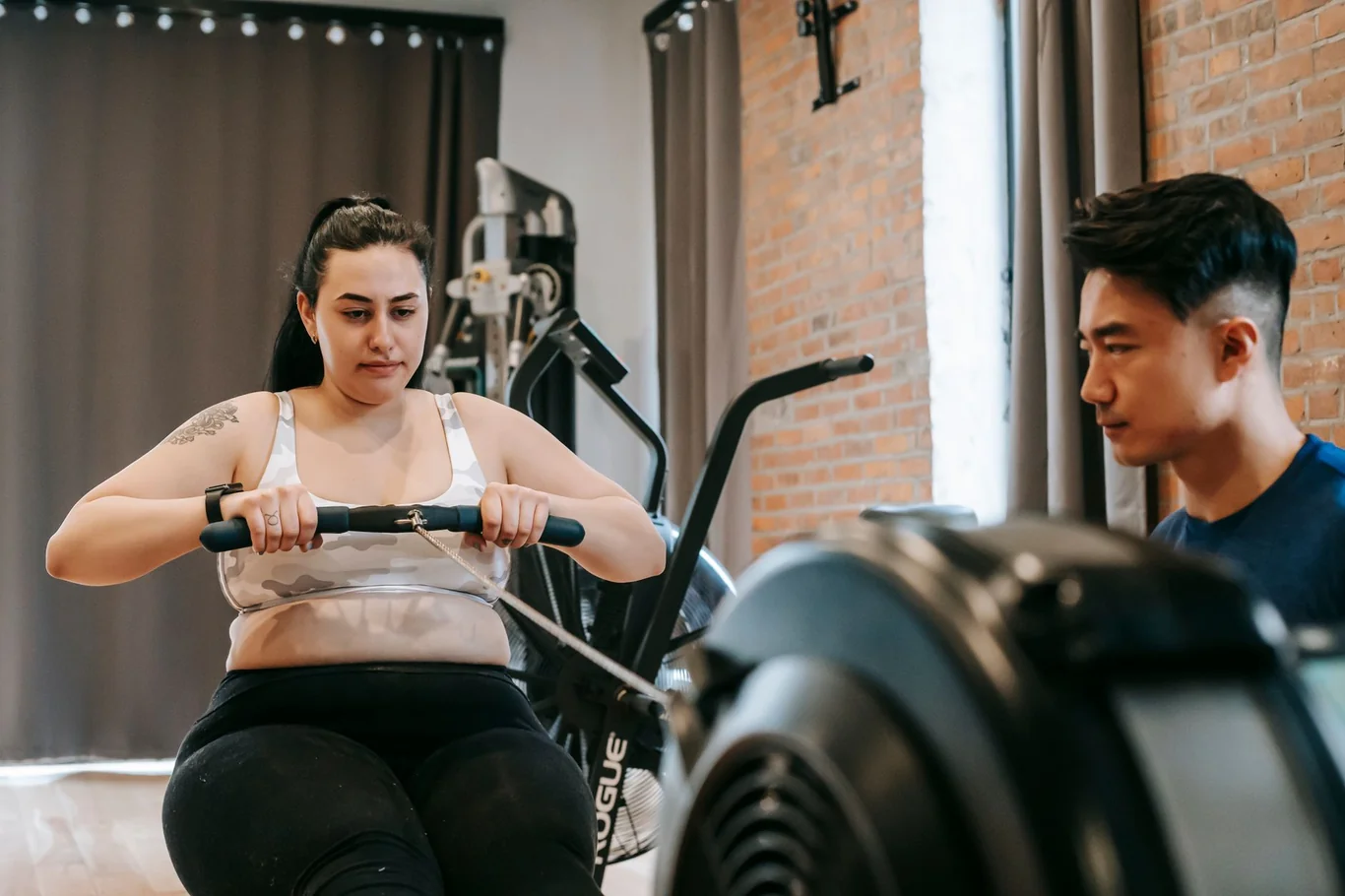 Woman rowing on a machine in a brick-walled gym while a trainer watches nearby