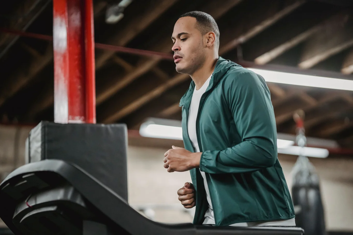 Man in a green jacket running on a treadmill in a gym with a punching bag nearby