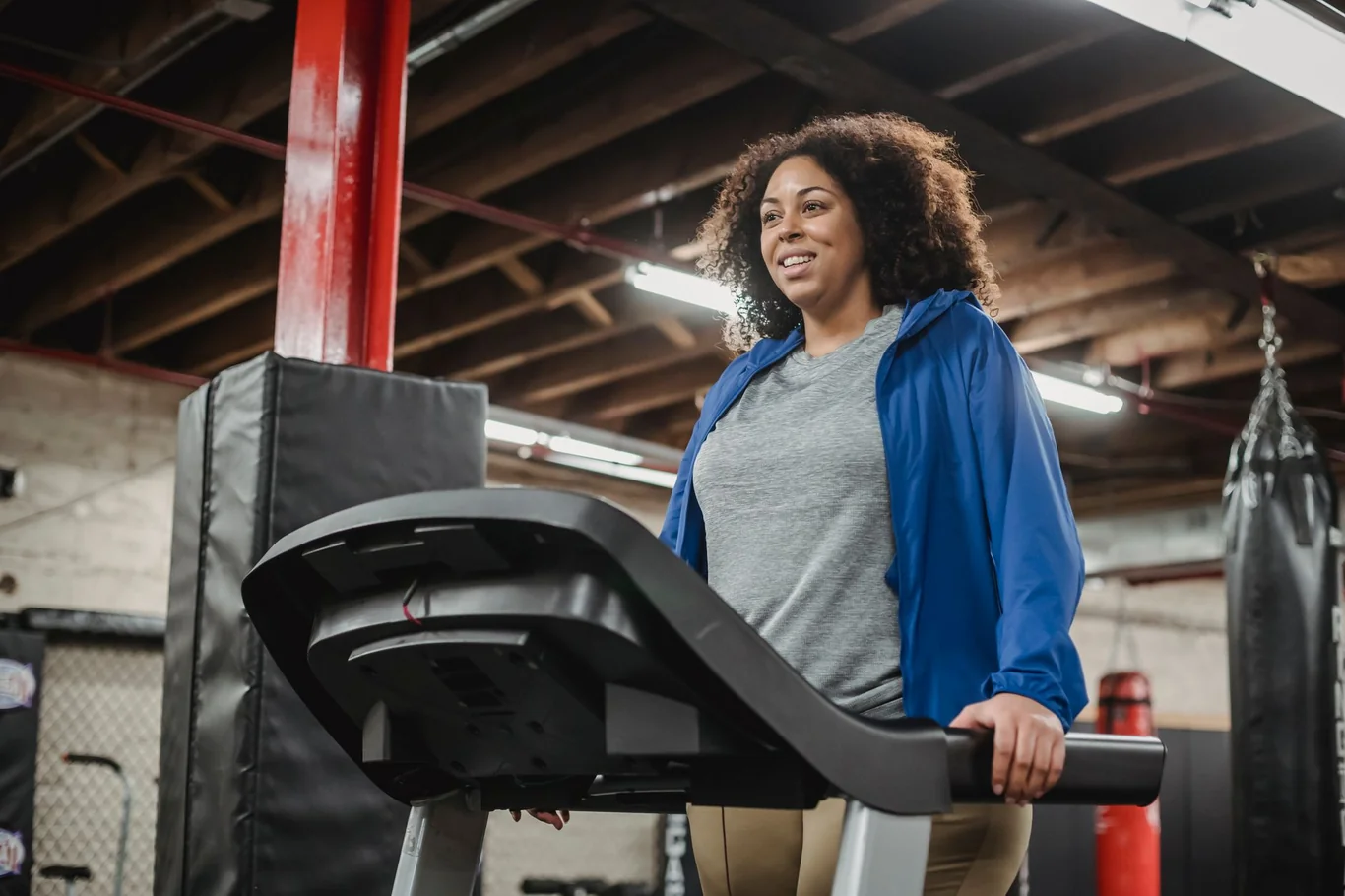 Woman in a blue jacket walking on a treadmill inside a gym with punching bags nearby