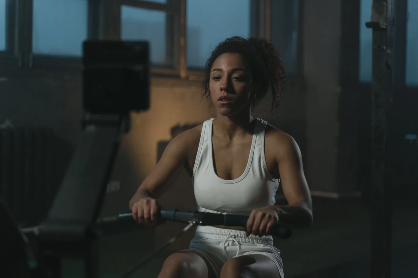 Woman in a white tank top gripping the handle of an indoor rowing machine