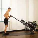 Man standing beside the Sunny Obsidian Surge water rower, upright storage position