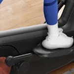 Close-up of a foot on the Merach elliptical pedal with the wood-finish flywheel visible
