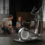 Man and woman kneeling beside the HARISON elliptical during setup, holding a pedal