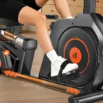 pooboo recumbent bike close-up of the seat rail adjustment lever, textured pedal with foot strap, and orange-ringed flywheel, with a rider's leg in position