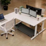 UREVO walking pad tucked under an L-shaped office desk with dual monitors
