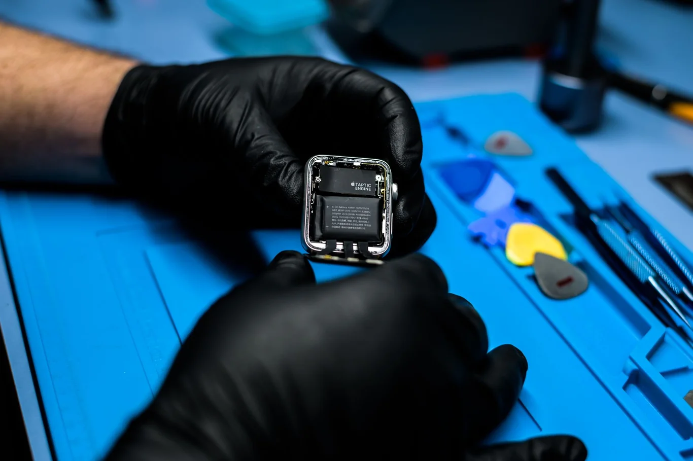 Technician in black gloves inspecting disassembled Apple Watch internals on a repair mat with tools nearby