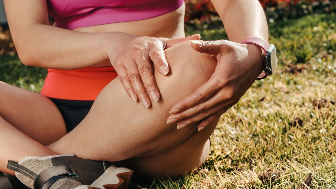 Female runner in workout clothes holding her knee while sitting on grass outdoors