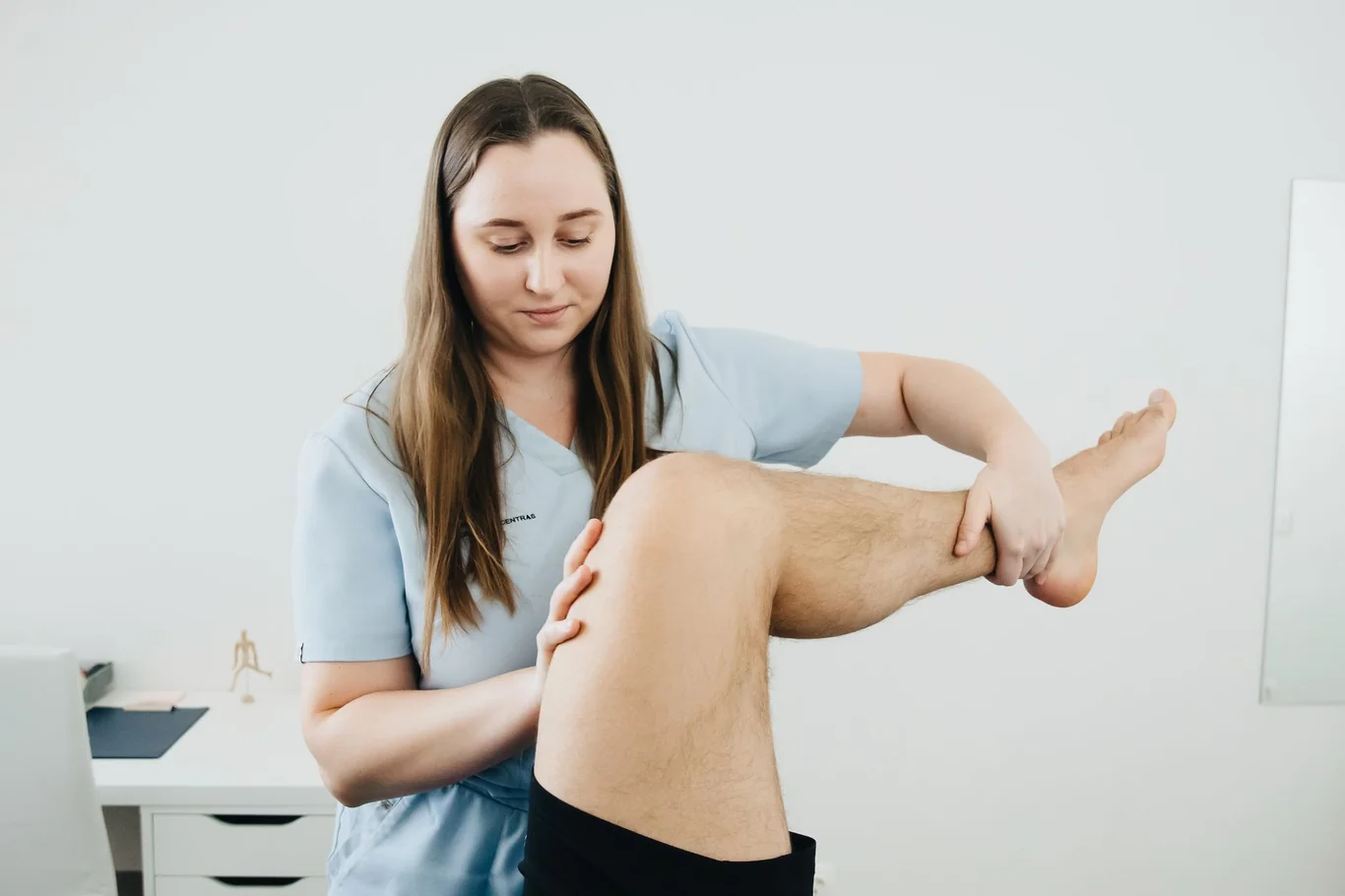 Physical therapist in blue scrubs manually examining a patient's knee during a clinic session
