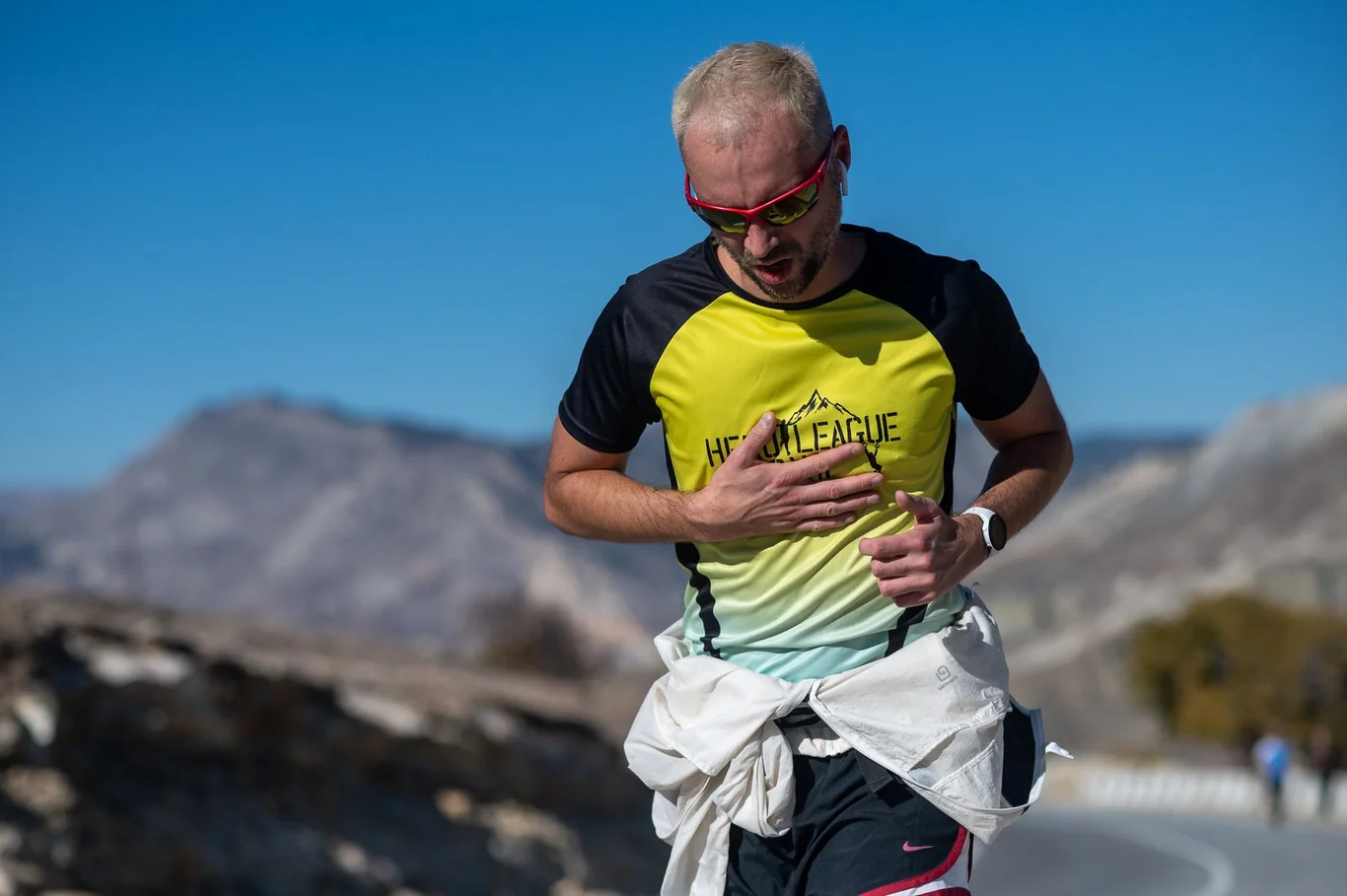 Man running at high intensity along a mountain road with hand on chest