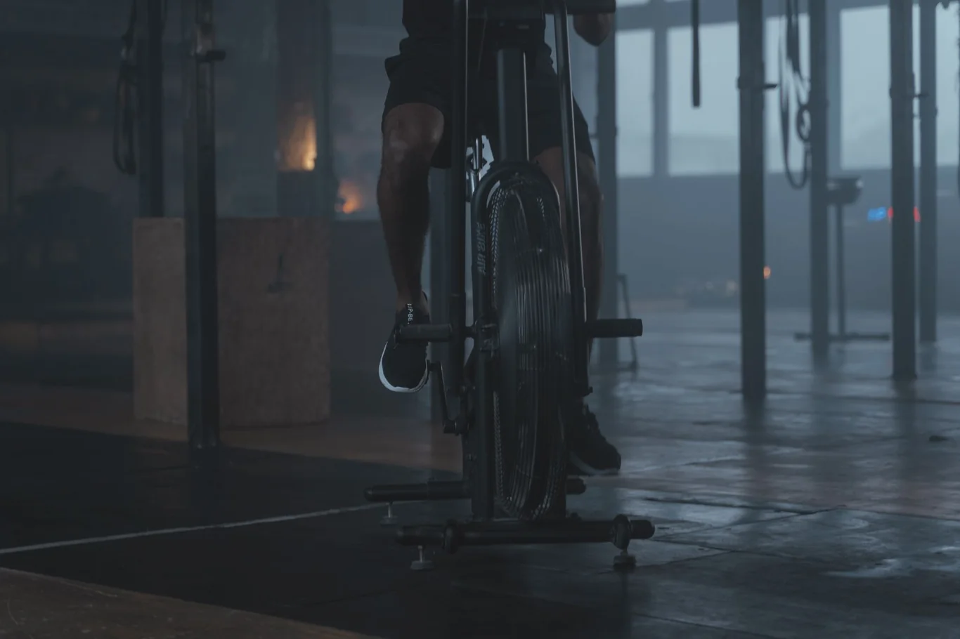 Close-up of a person's lower legs pedaling on an air bike in a dark gym