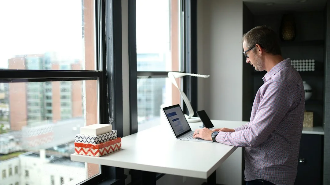 Man working at a standing desk by large windows overlooking a city