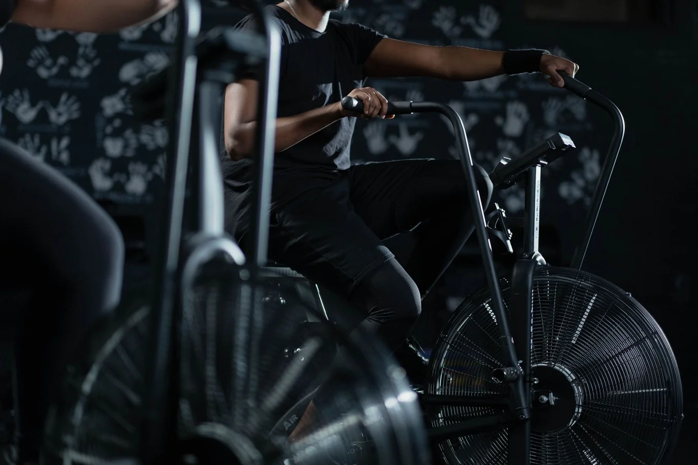 Man pedaling an air bike in a dark gym, gripping handlebars in an upright riding position