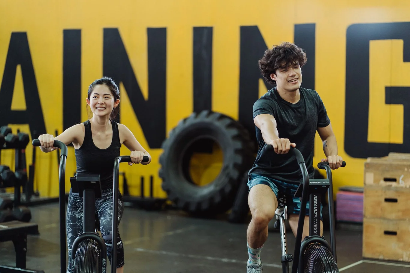 Man and woman smiling while pedaling air bikes side by side in a gym