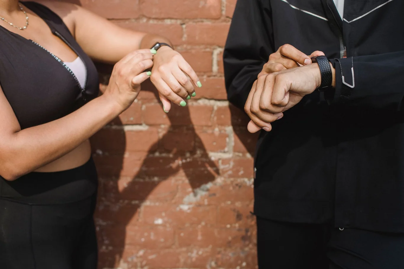 Two people comparing fitness bands on their wrists outdoors against a brick wall
