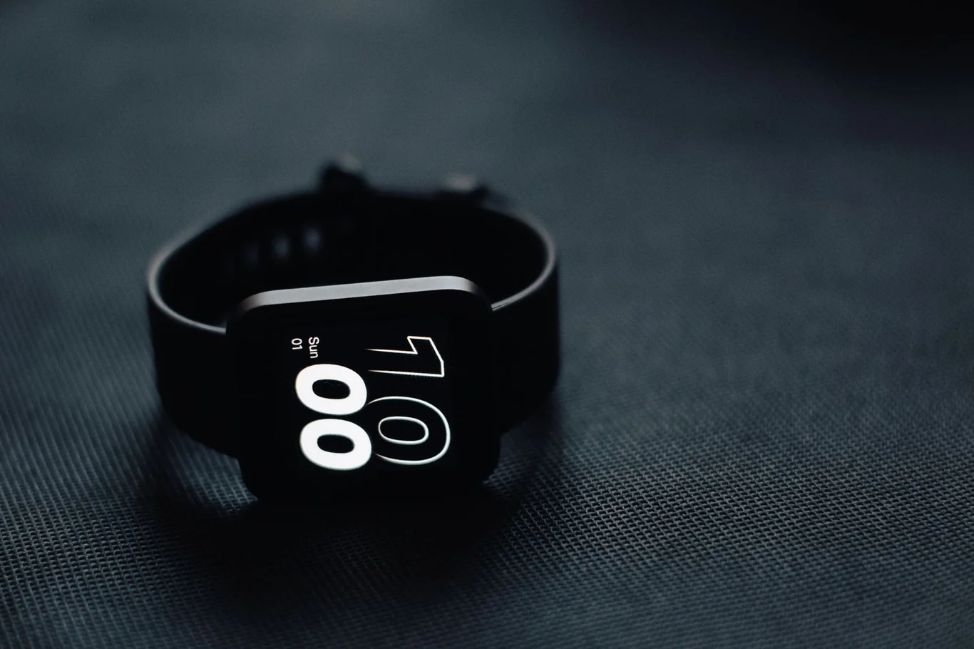 Black square smartwatch with digital clock face on dark fabric surface