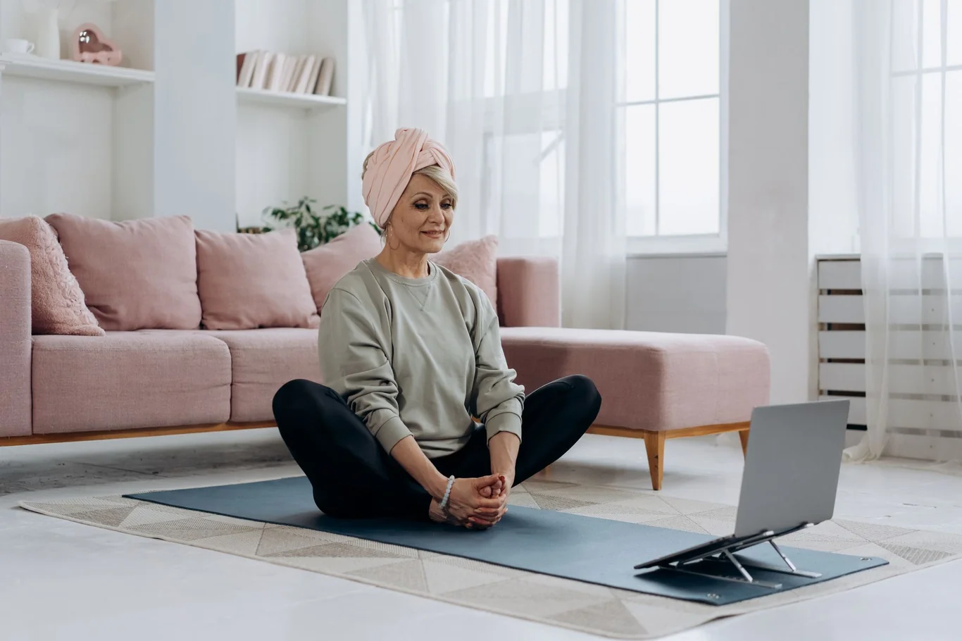 Woman sitting on a yoga mat in a living room following a workout class on a laptop screen