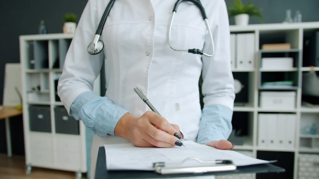 Doctor in white coat writing on a clipboard with a stethoscope around the neck in a medical office