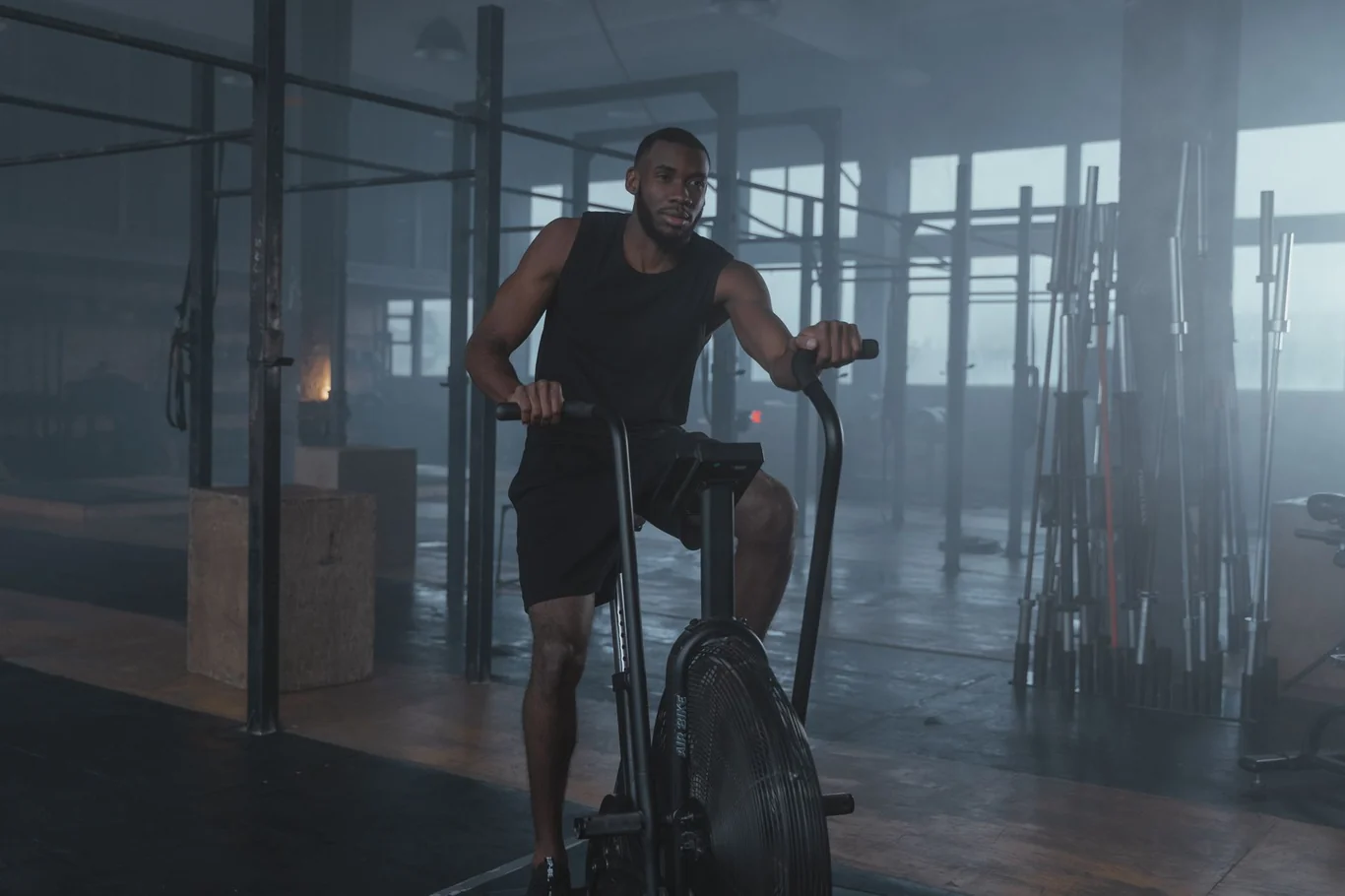 Man in black workout clothes pedaling an air bike in a dark industrial gym