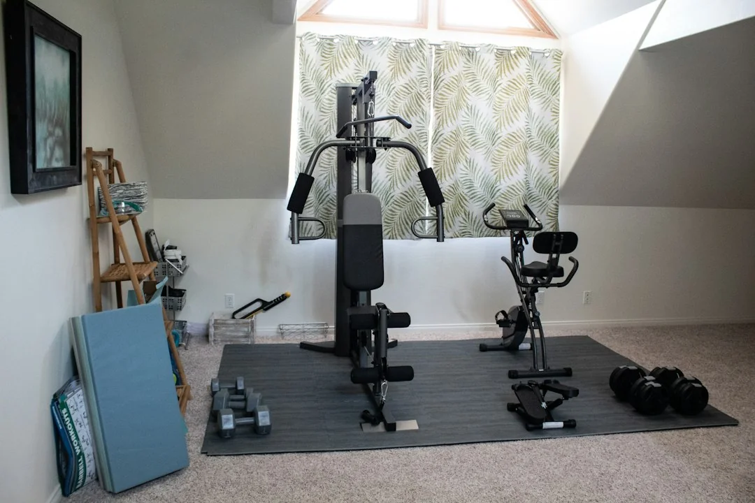 Home gym setup in an attic room with a cable machine, exercise bike, dumbbells, and a rubber floor mat under natural window