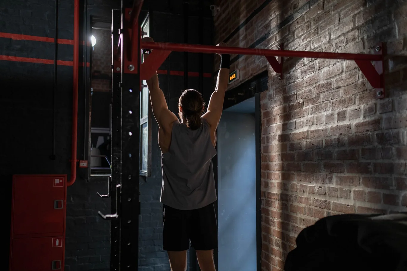 Man performing a pull-up on a red pull-up bar in a dark indoor gym