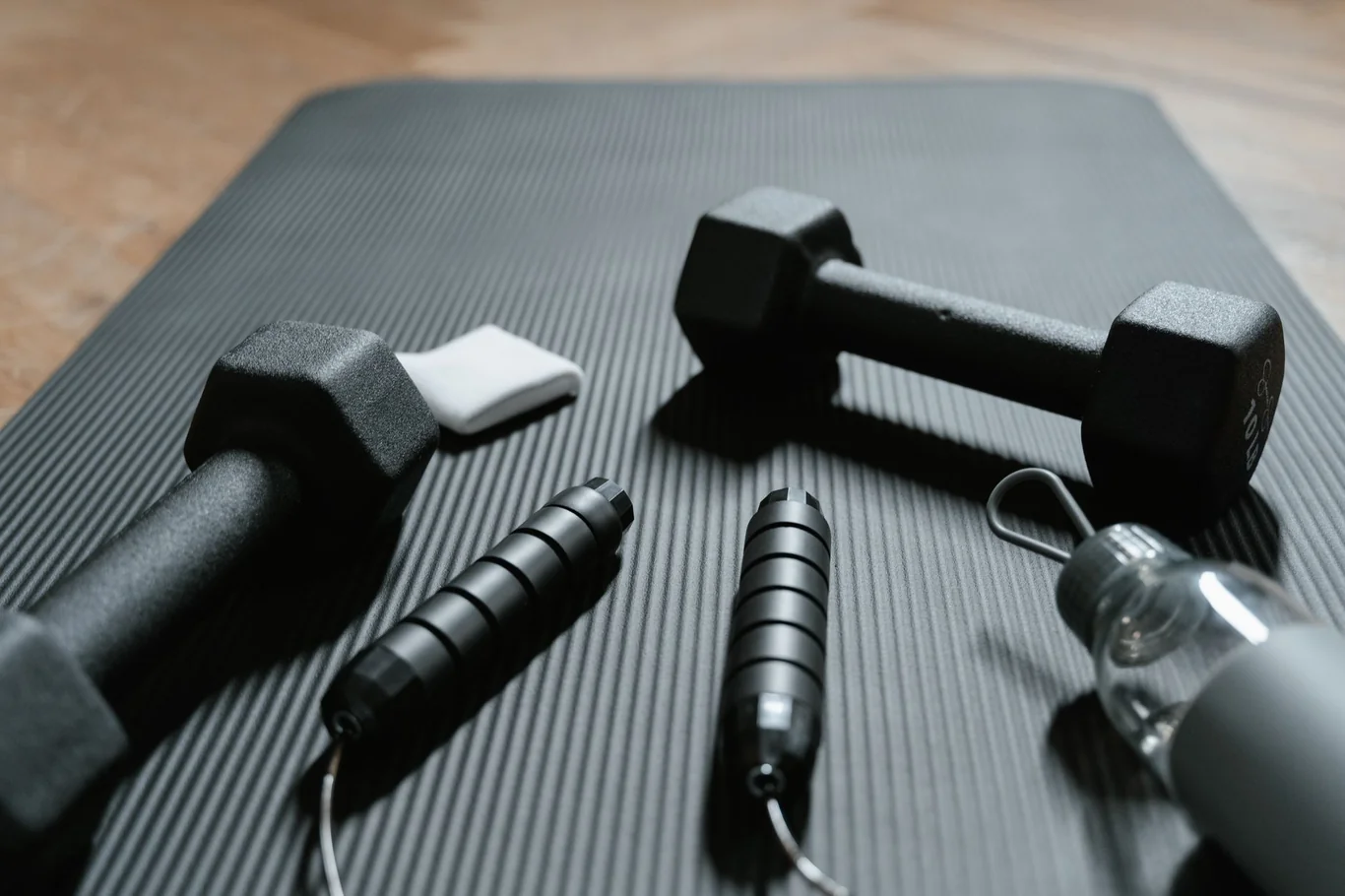 Black hex dumbbells and jump rope laid out on a dark exercise mat at home