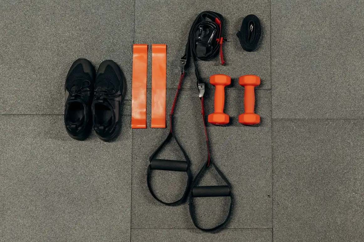 Compact home gym equipment flat lay with orange dumbbells, resistance bands, TRX straps, and jump rope on rubber floor