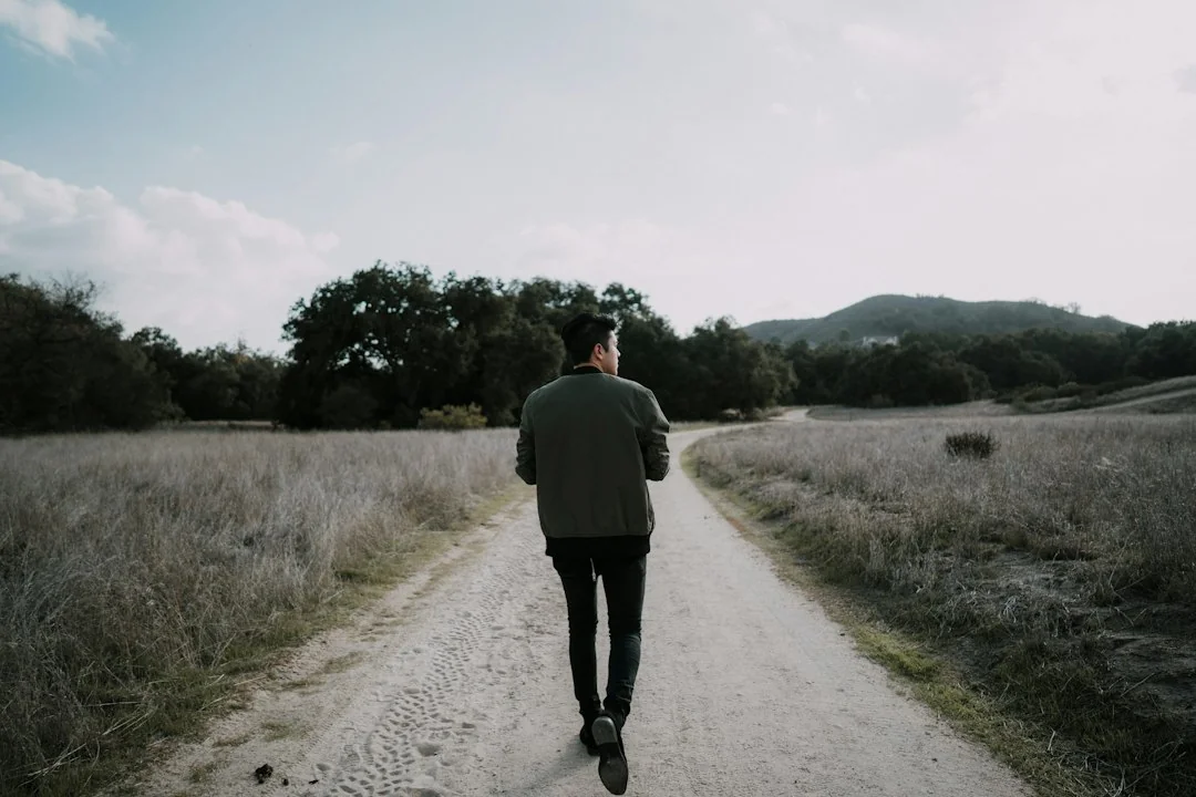Man walking alone on a dirt path through open fields toward tree-covered hills