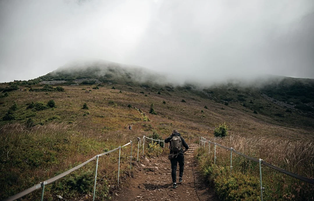Hiker with backpack walking up a misty mountain trail toward cloud-covered peak