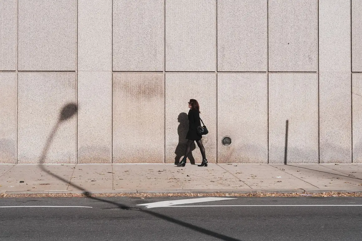 Woman walking alone along a wide sidewalk beside a stone building wall