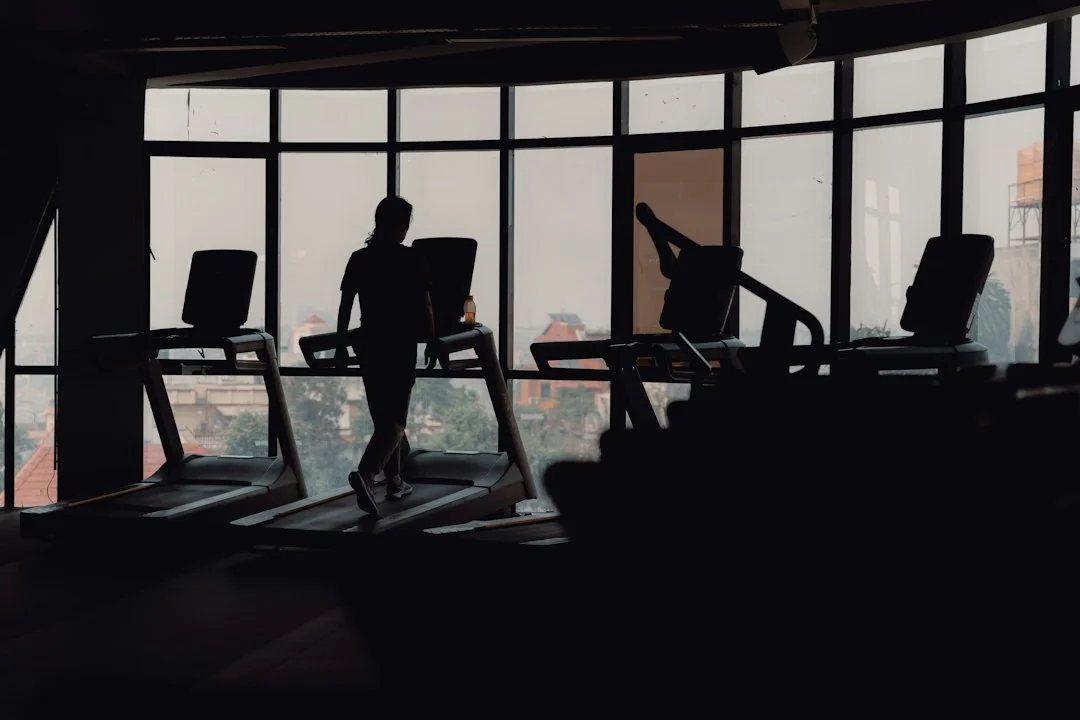 Person walking on a treadmill in a dark gym with large windows in the background