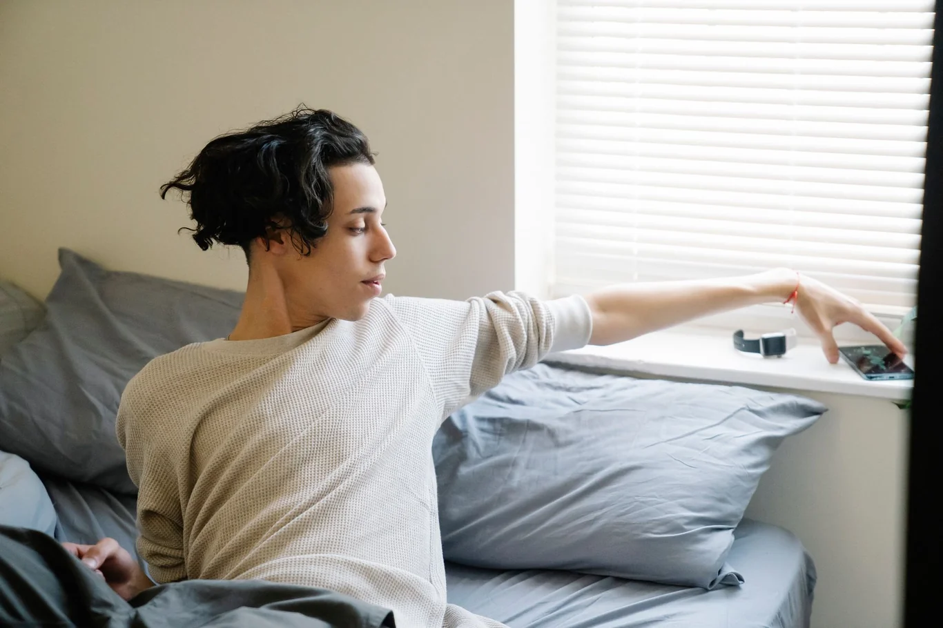 Woman sitting in bed reaching for her phone on a windowsill in the morning