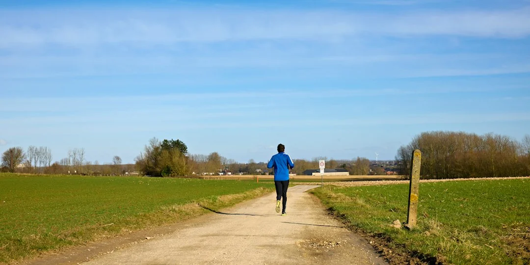 Runner in blue shirt jogging alone on a gravel path through open fields under a blue sky