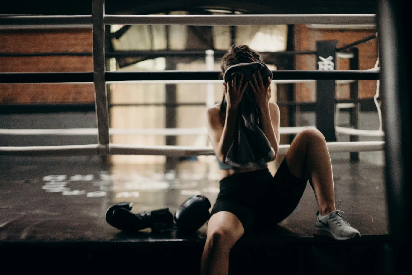 Female athlete sitting on a boxing ring floor, covering her face with a towel after a hard workout