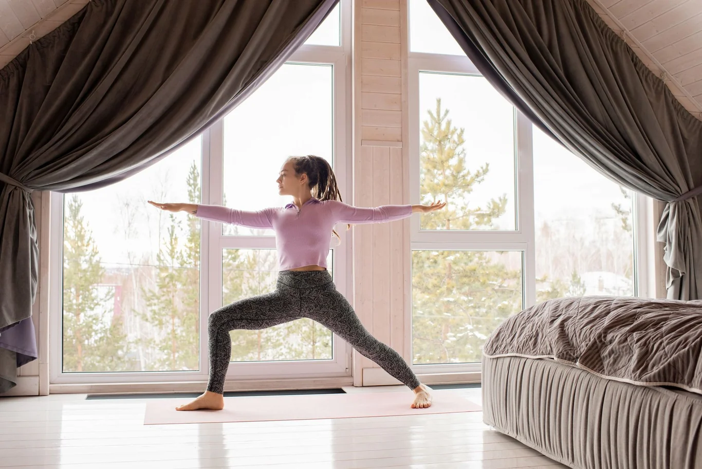 Woman holding a warrior yoga pose on a mat beside a large window