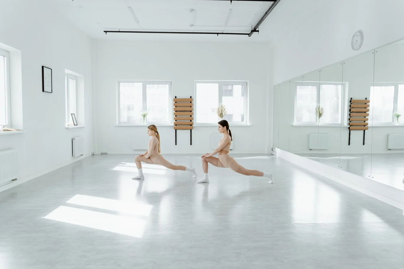 Two women doing lunge stretches in a bright studio with a full-length wall mirror