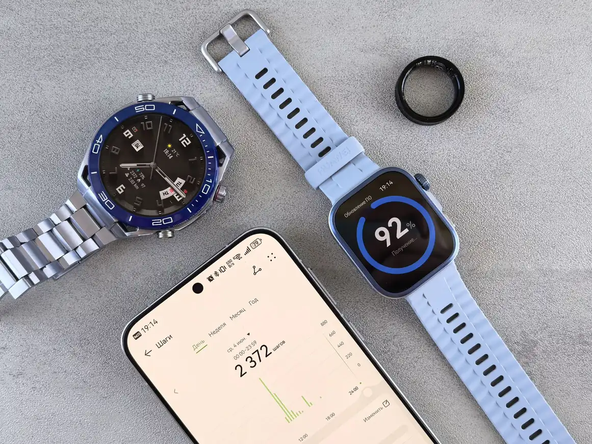 Two smartwatches and a smart ring laid next to a smartphone displaying fitness tracking data on a gray surface