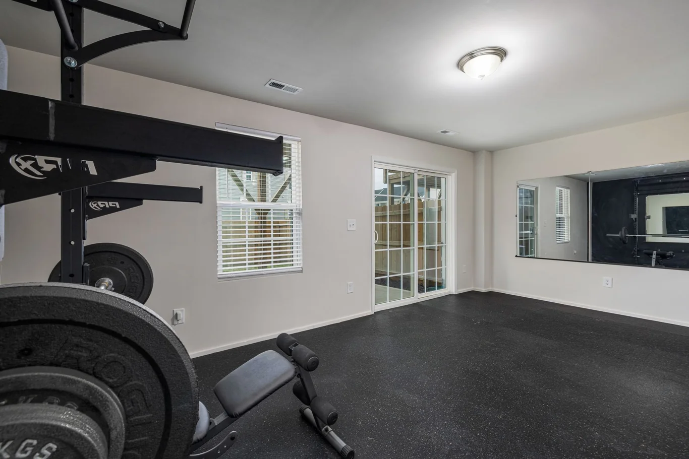Empty home gym corner with a weight rack, bench, and stacked plates on black rubber flooring