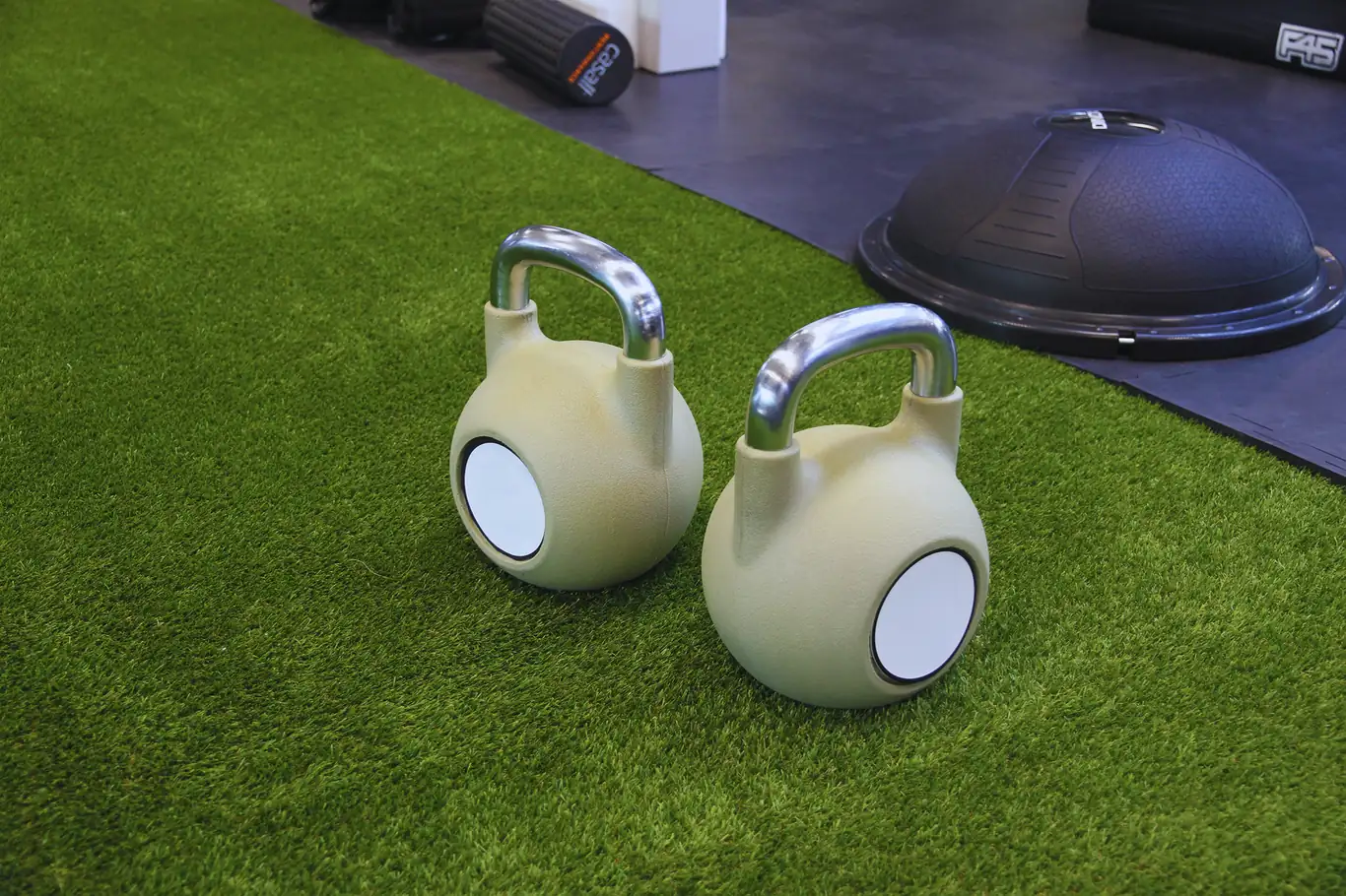 Two kettlebells sitting on green turf flooring near workout equipment
