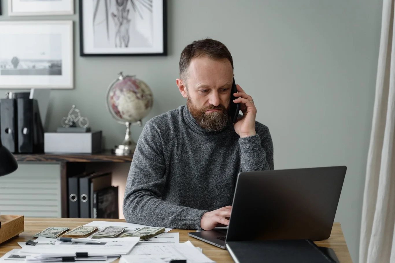 Man on a phone call while checking his laptop at a home desk with cash nearby
